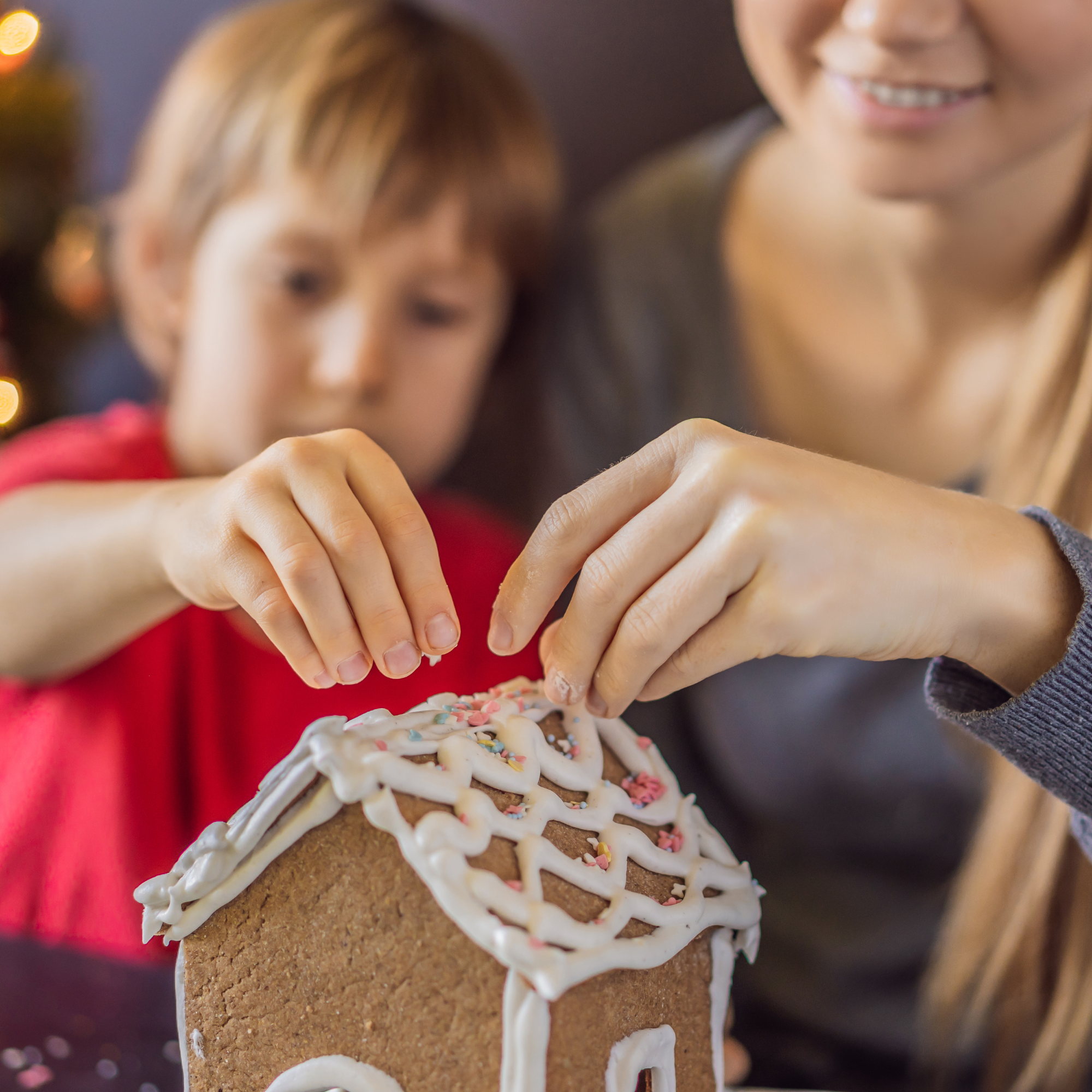 A woman and child decorate a gingerbread house with icing and sprinkles. Their hands gently place decorations, conveying a warm, festive atmosphere.