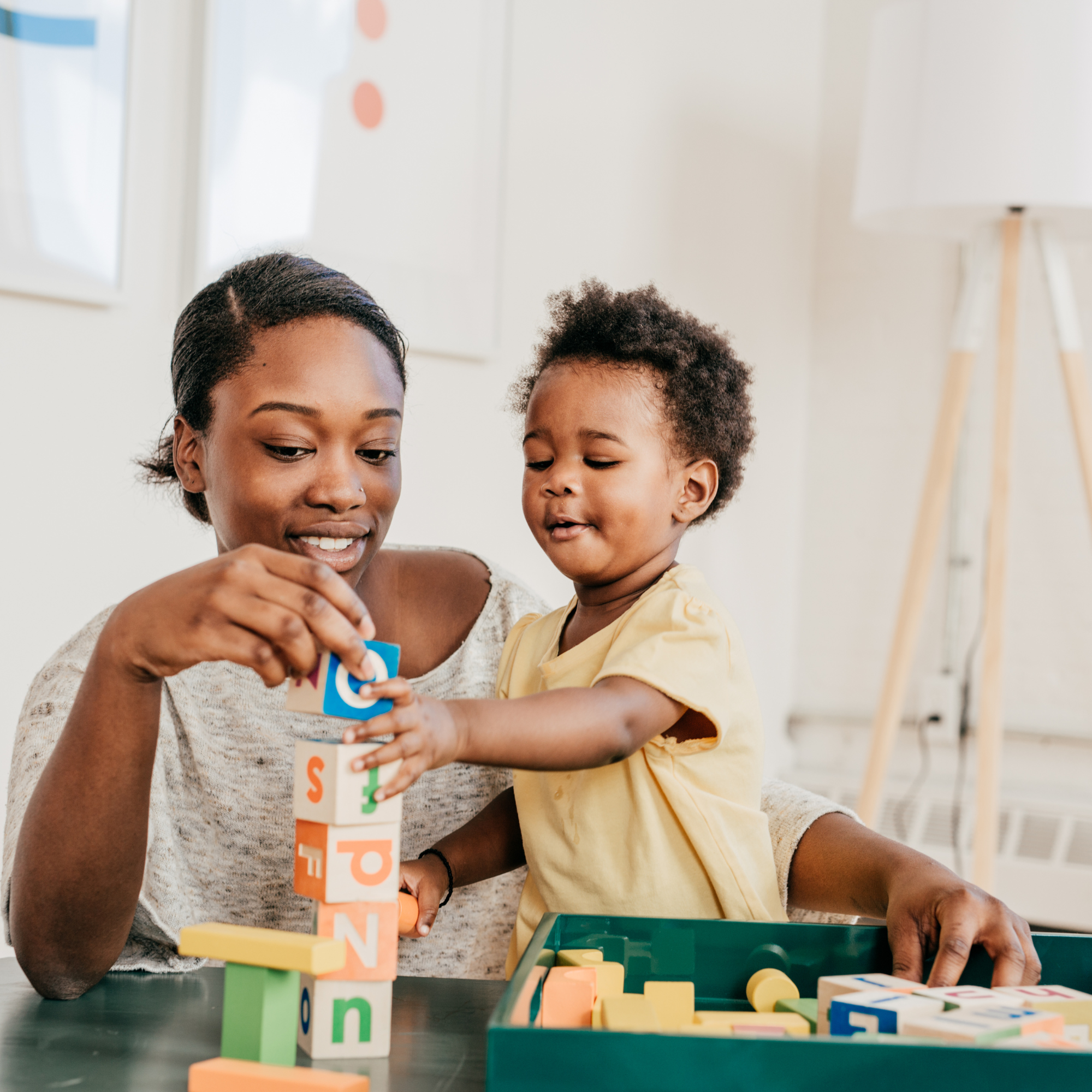 A mother and toddler play with colorful alphabet blocks, stacking them on a table in a bright room, conveying a sense of learning and possible early intervention.