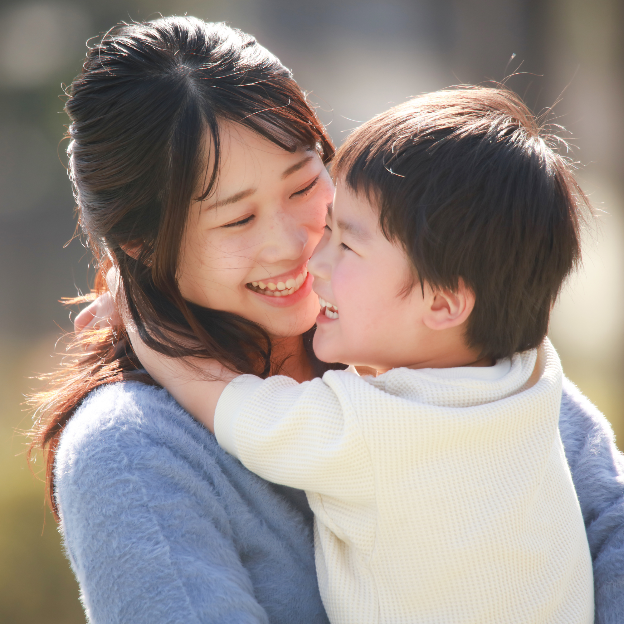 A mother and a child share a joyful embrace outdoors. Both are smiling brightly, capturing a warm, affectionate moment in natural lighting.