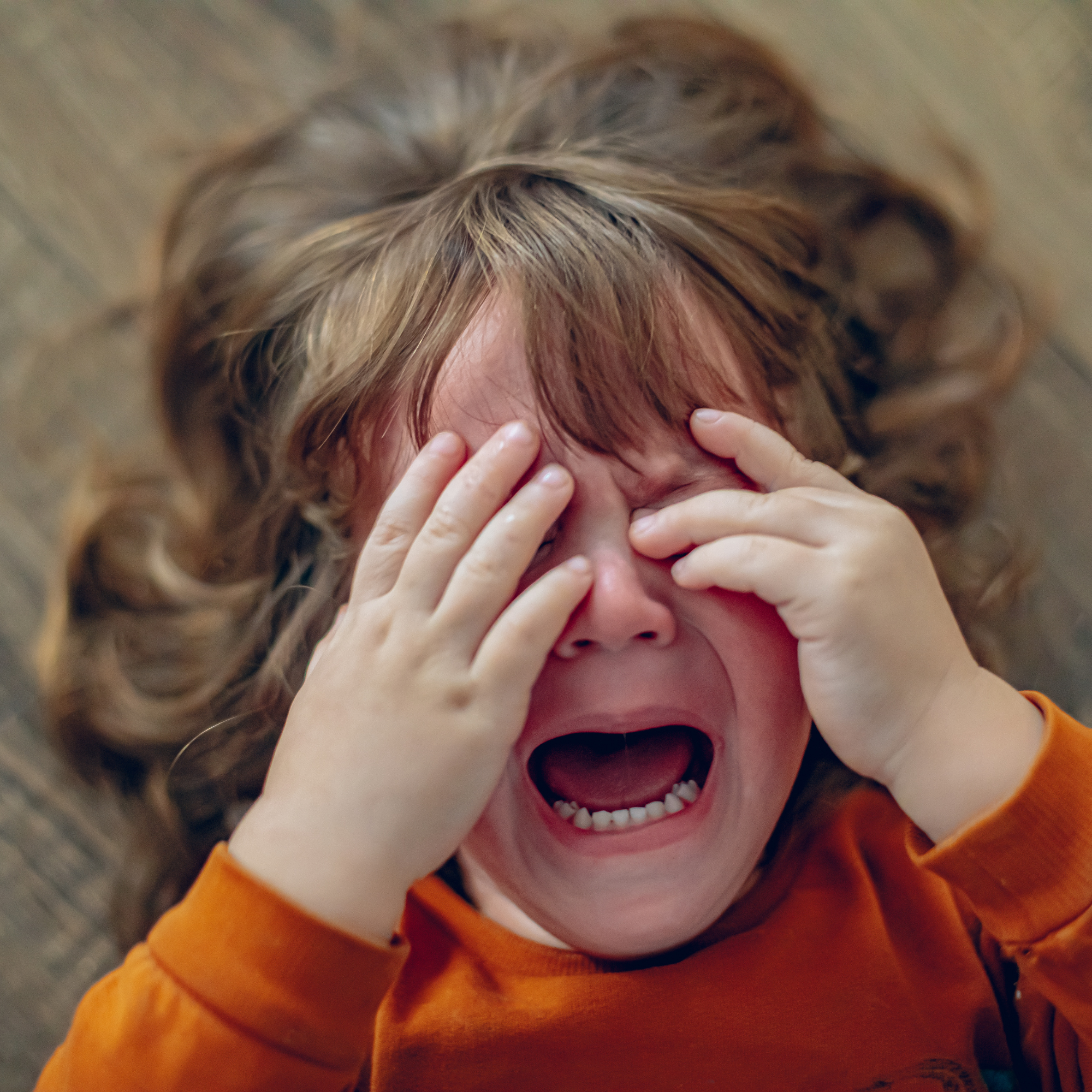 A young child lying on the floor, wearing an orange shirt, is crying with hands covering their eyes and a wide-open mouth, conveying distress and sadness.