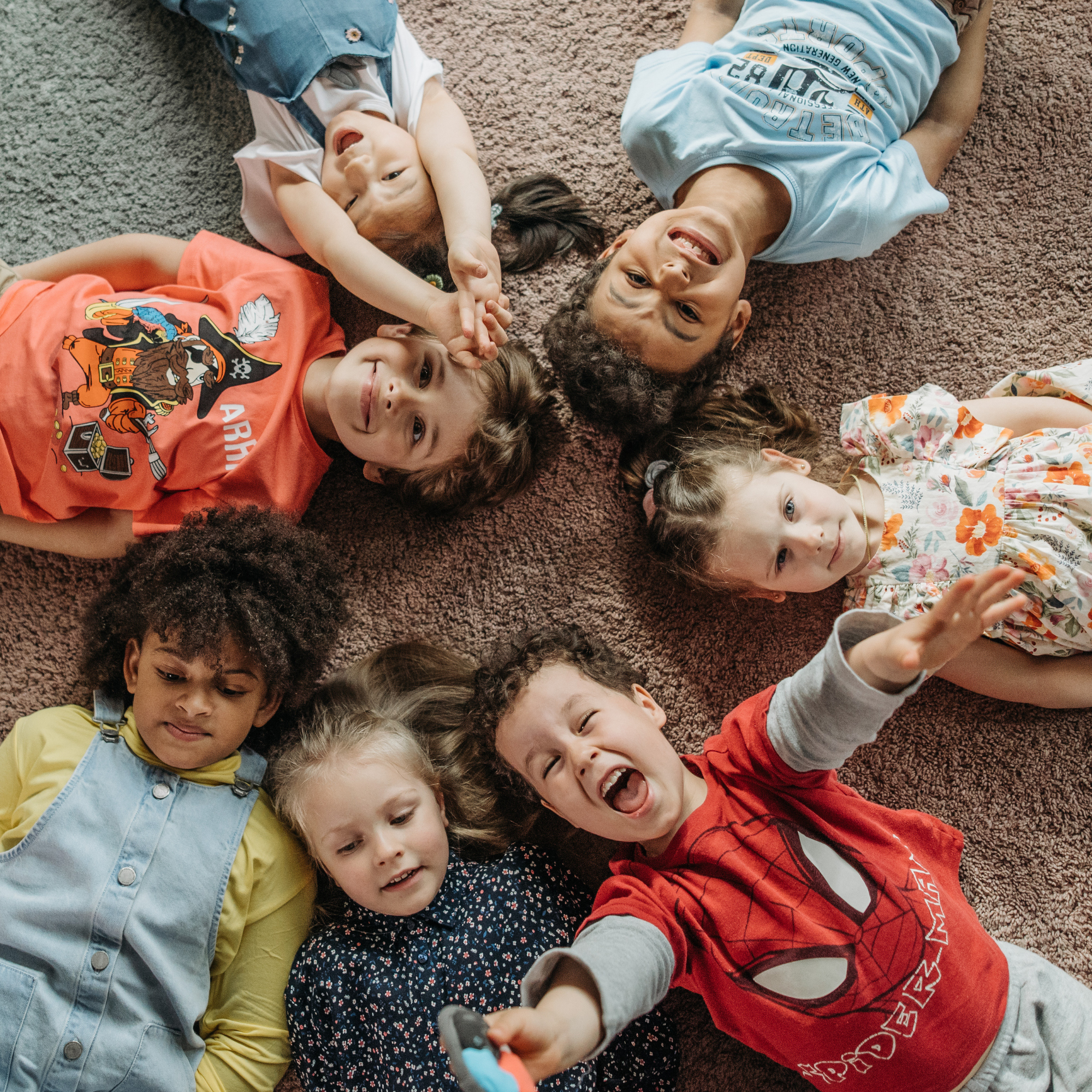 A group of seven children, lying on a carpet in a circular formation, smiling and laughing. Their heads meet at the center, creating a joyful scene.