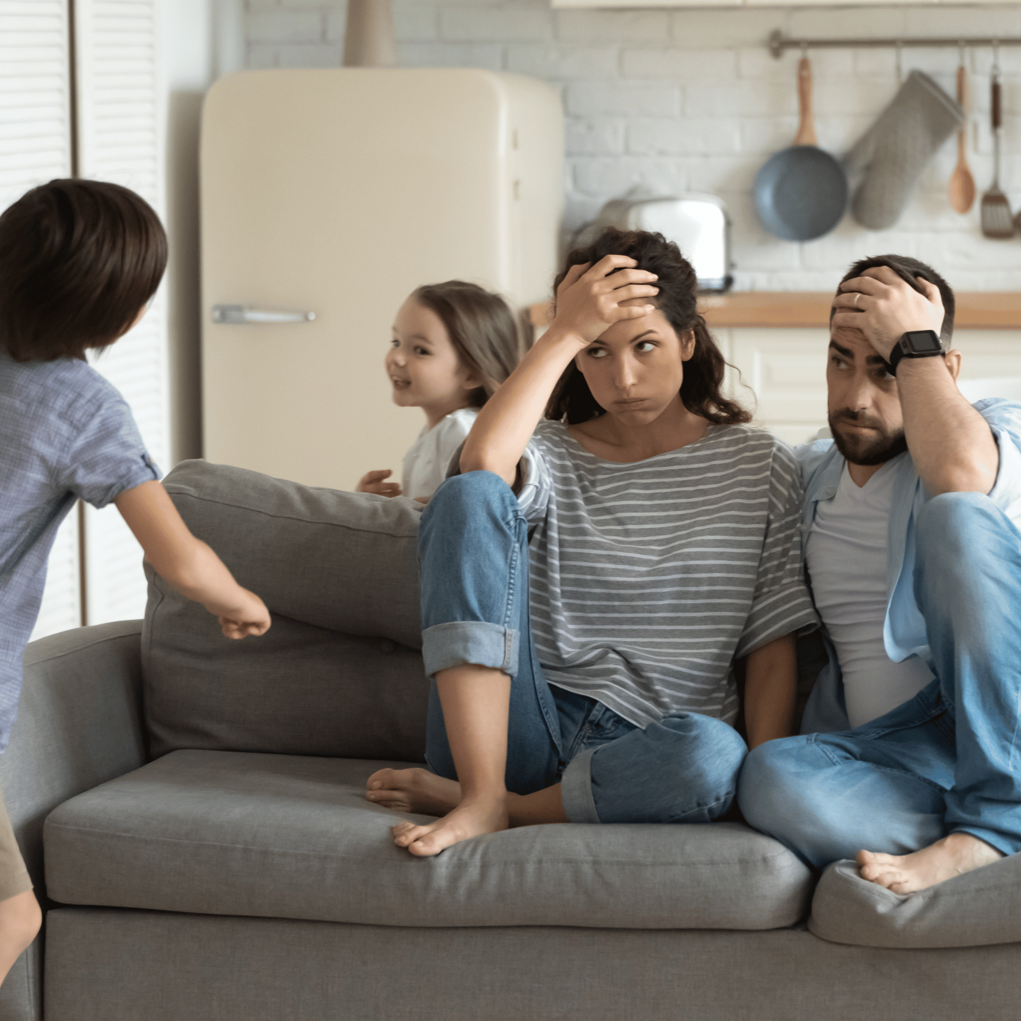 Parents sitting on a couch look exasperated as two energetic children play around them in a cozy kitchen setting. The mood is chaotic yet humorous.