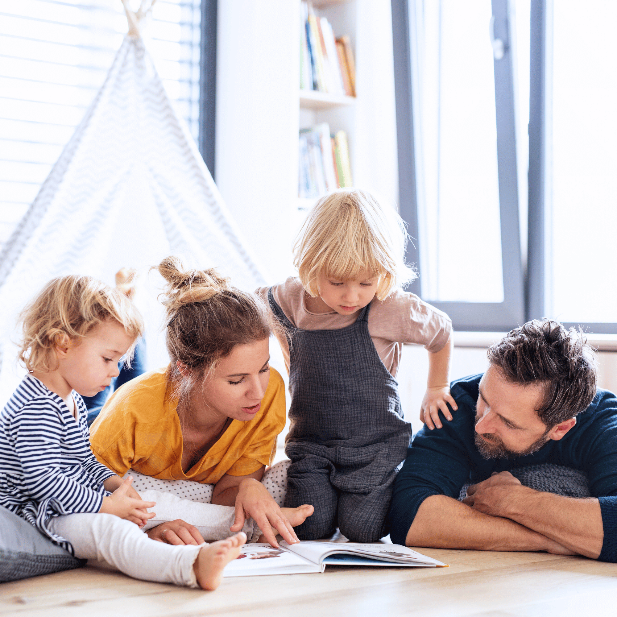 A family of four, including two young children, enjoys reading a book together on the floor. The mood is warm and focused, with light streaming in.