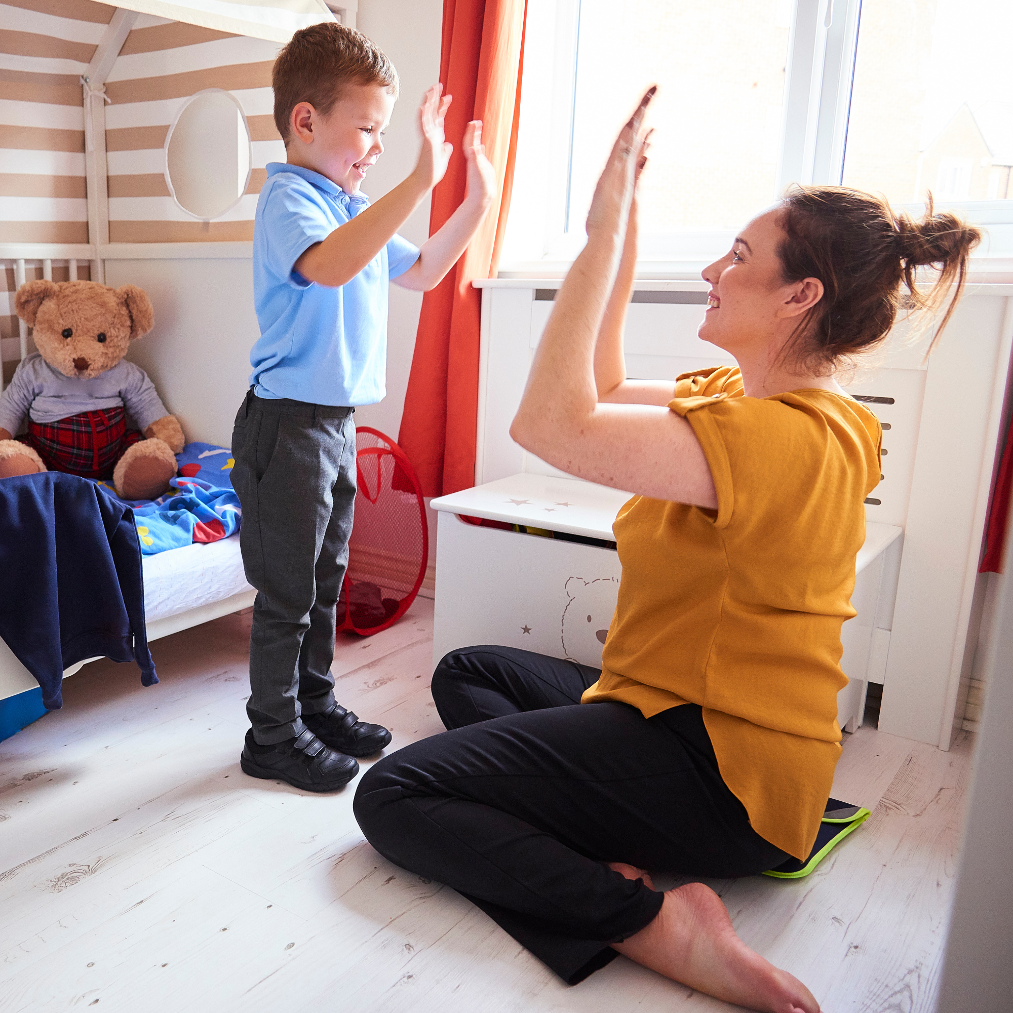 A joyful woman sitting on the floor high-fives a smiling child in a bedroom. A teddy bear and toy-filled bed are visible. Bright, cheerful atmosphere.