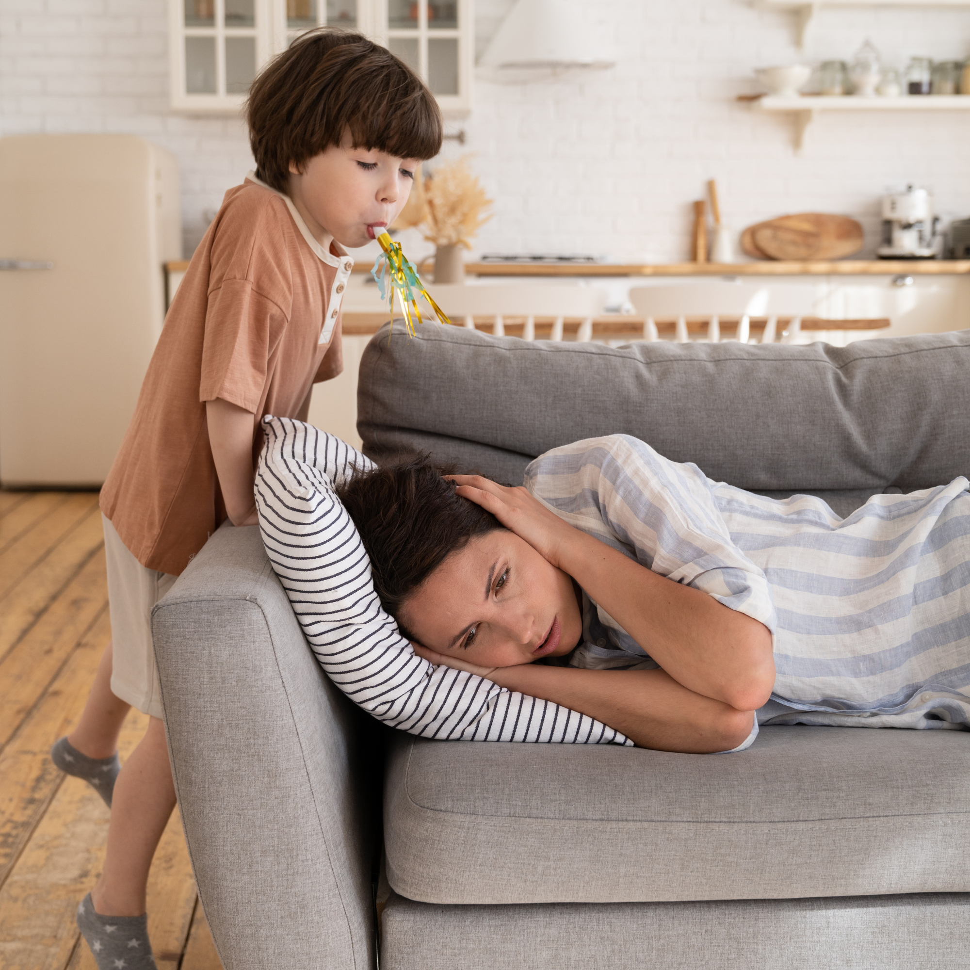 A child playfully blows a party horn next to a woman lying on a gray couch, looking tired and covering her ears. The setting is a cozy, sunlit living room.