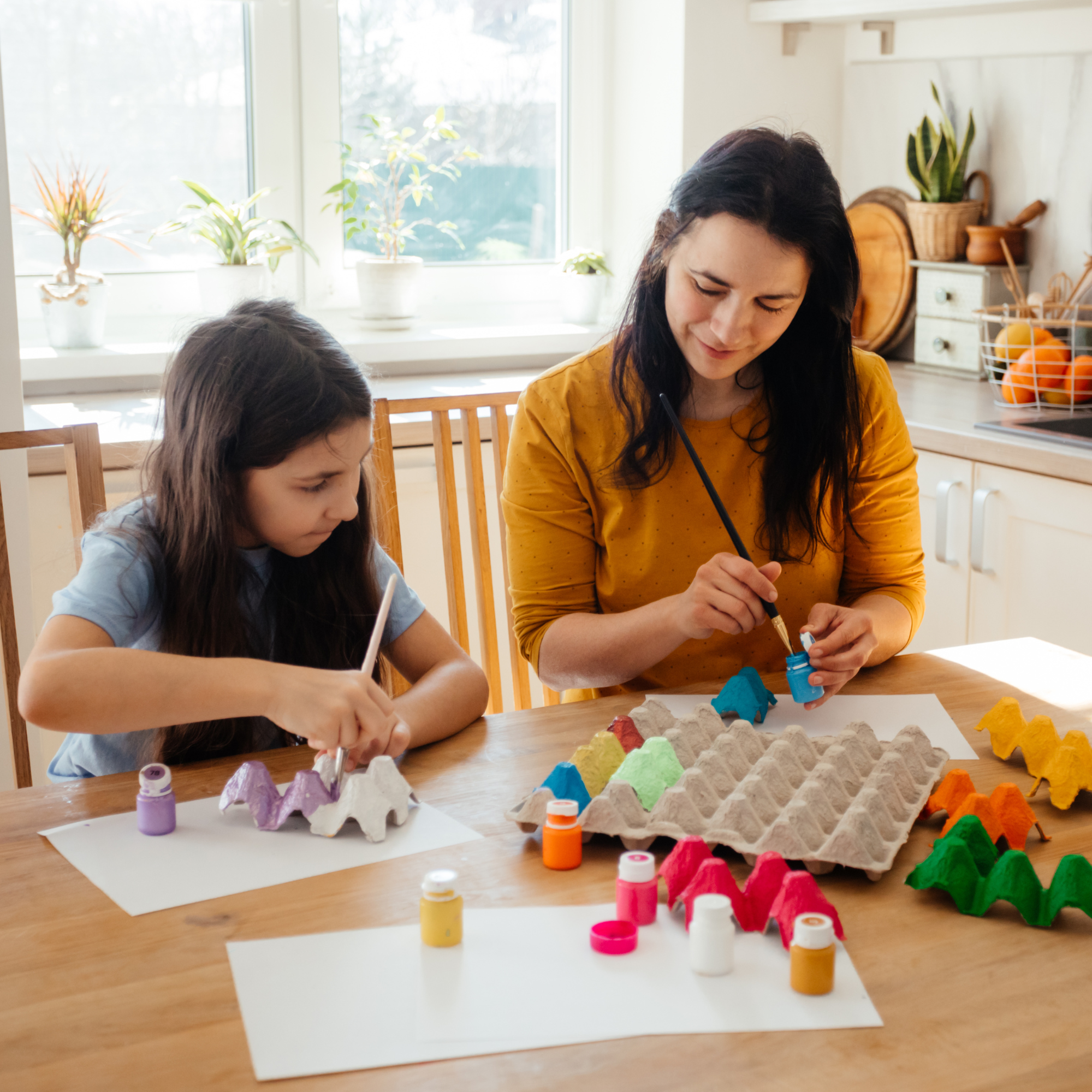 A woman and a child are painting egg cartons with bright colors at a wooden table. Sunlight streams through a window, creating a warm, creative atmosphere.