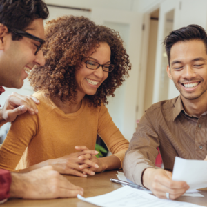 Three people smiling and reviewing documents at a table, conveying a sense of teamwork and positivity. They appear to be in a well-lit office.