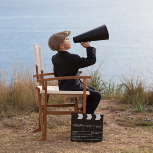 Young boy in director's chair by a lake, wearing a suit and cap, holds a megaphone. Clapperboard on ground; scene suggests imaginative play.