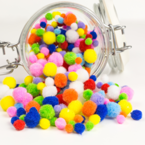 A glass jar tipped over with colorful pom-poms spilling out. The pom-poms are vibrant in various sizes, creating a playful, cheerful scene.