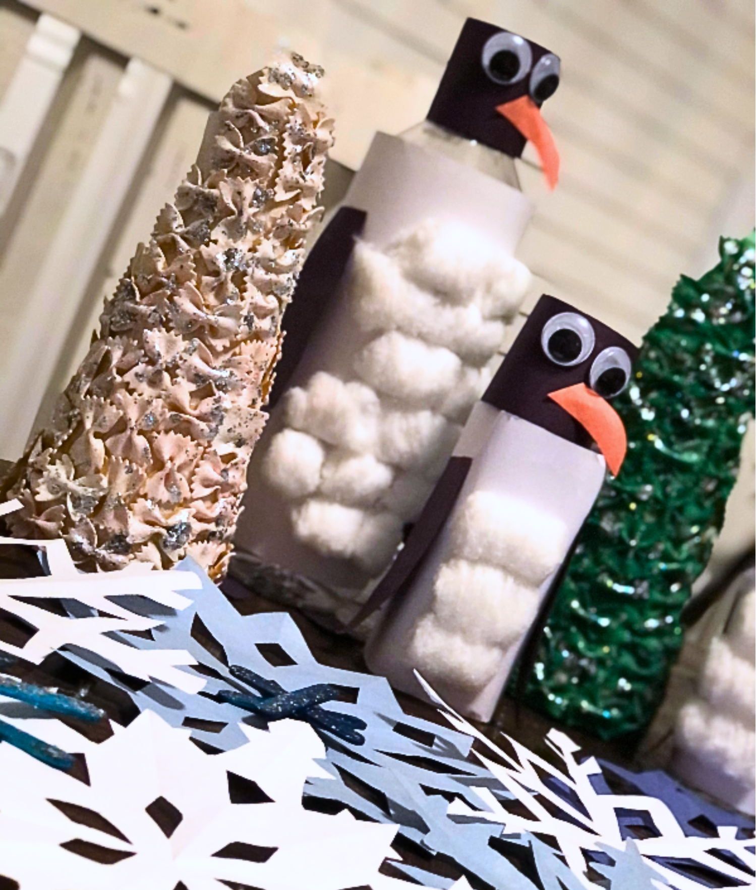 Festive table with handmade crafts: two penguins made of bottles and cotton, surrounded by paper snowflakes and pasta trees, conveying a winter vibe.