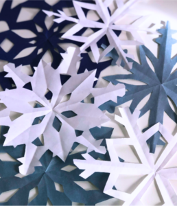 Paper snowflakes in shades of blue and white are layered on a light surface, showcasing intricate, symmetrical designs, evoking a wintry, festive atmosphere.