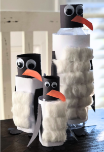 Three penguin crafts made from bottles, decorated with cotton balls, black paper wings, googly eyes, and orange beaks, creating a playful scene.