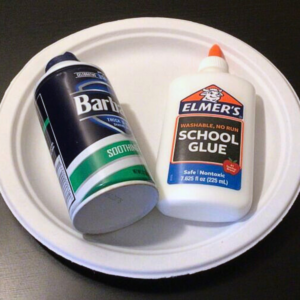 A can of Barbasol shaving cream and a bottle of Elmer's school glue sit on a white paper plate, placed on a dark surface.