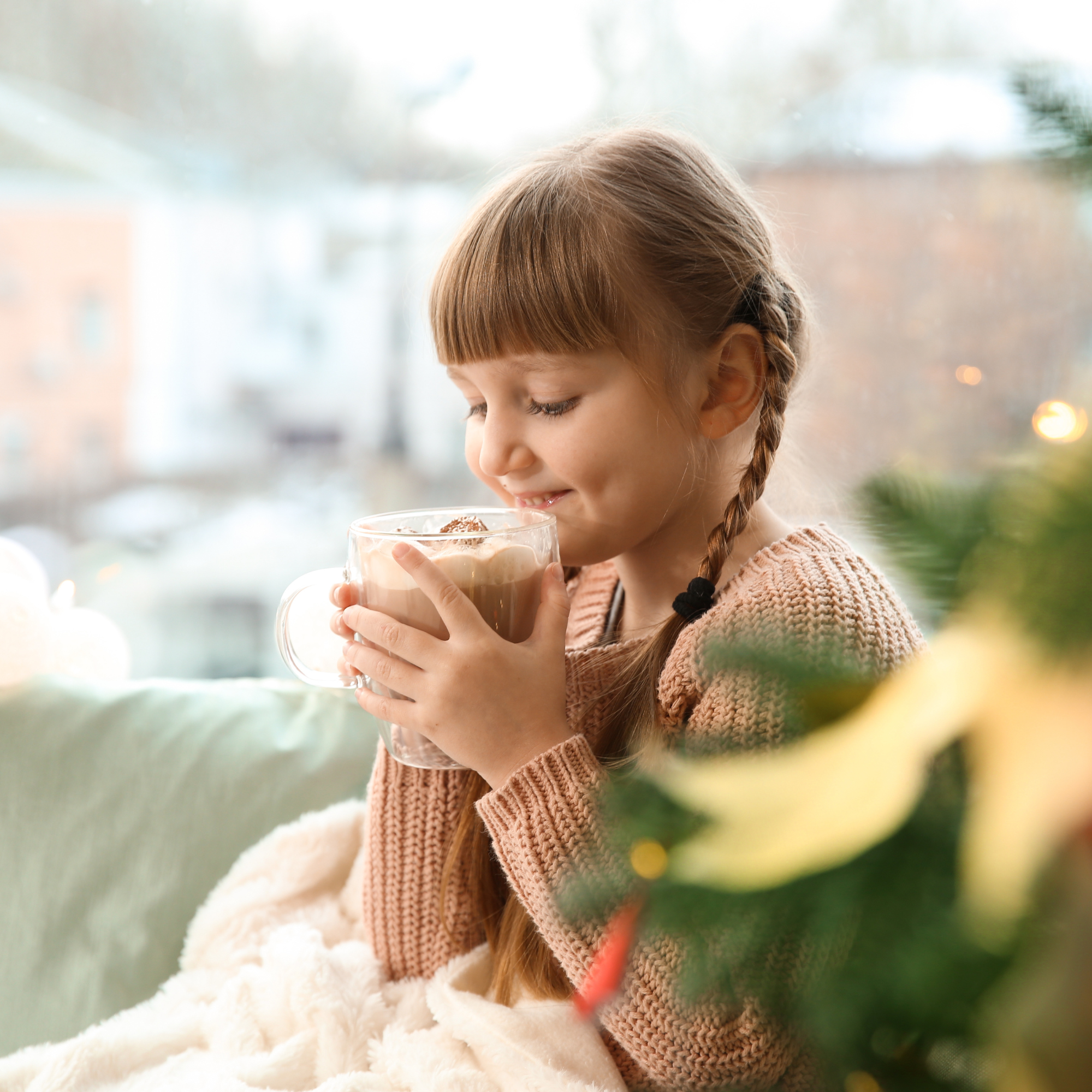 A young girl with braided hair smiles while holding a mug of hot chocolate, wrapped in a blanket by a window. A Christmas tree is in the foreground.