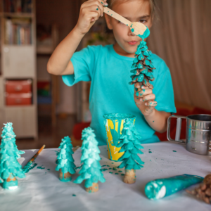 A child in a teal shirt paints pine cones turquoise, mimicking trees. The scene is colorful and creative, featuring brushes, paint, and a focused expression.