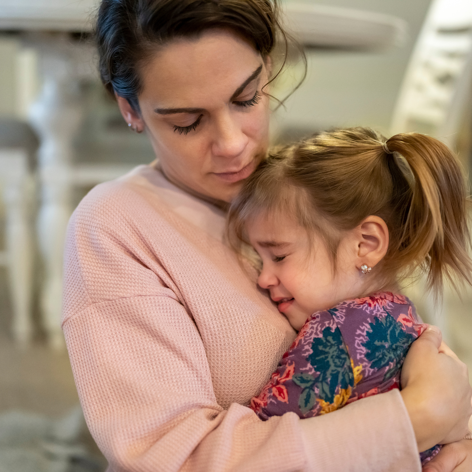 A woman in a pink sweater gently hugs a crying young girl with light brown hair in a ponytail. They share an emotional, comforting moment indoors.