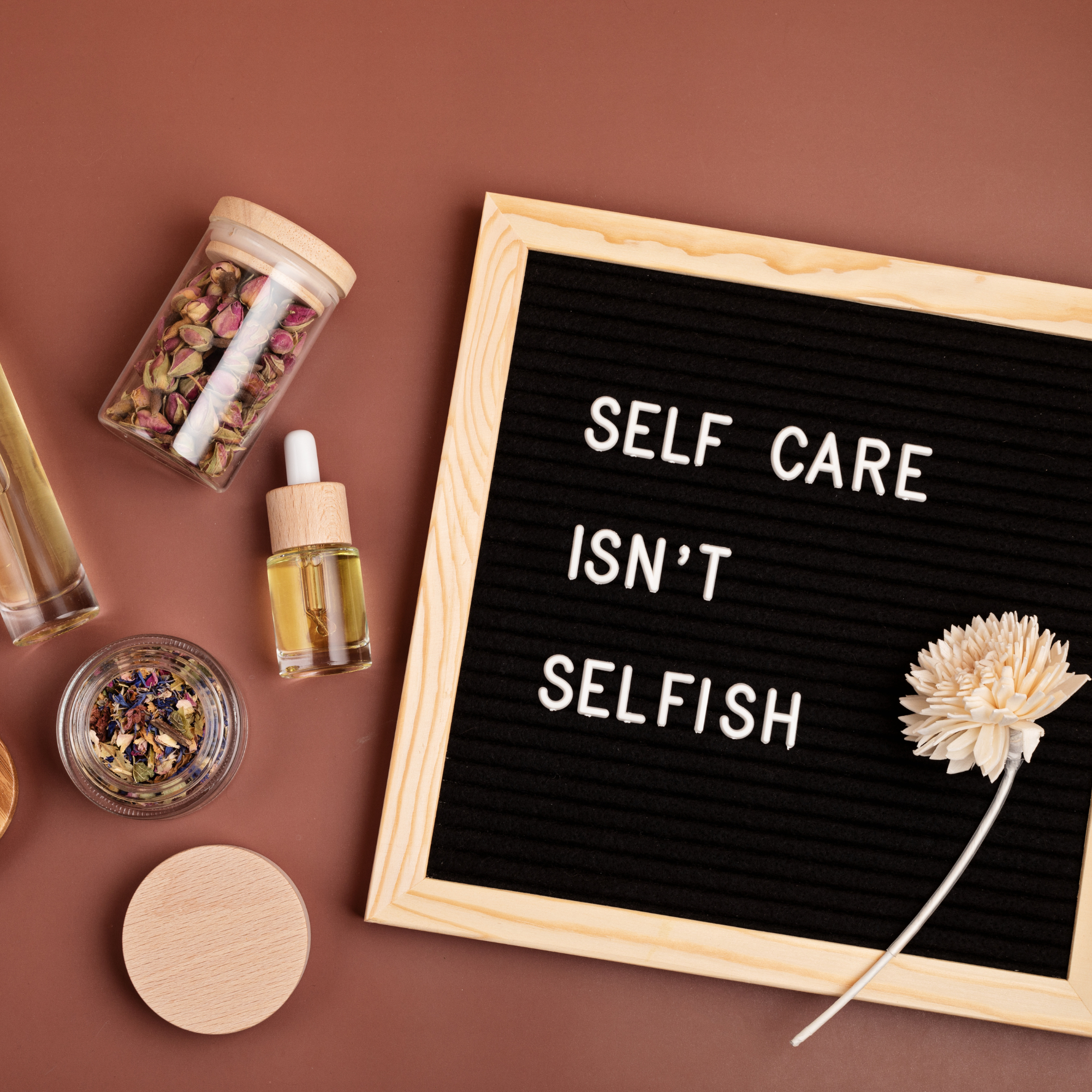 A wooden-framed sign on a brown background reads "Self Care Isn't Selfish." Surrounding it are bottles of essential oils and dried flowers, suggesting relaxation.