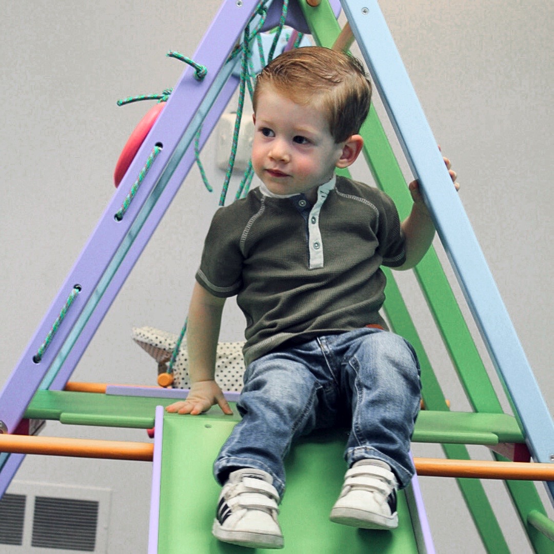 A young boy sits relaxed on a colorful indoor slide, with a playful expression. The slide features pastel-colored bars and ropes, creating a cheerful scene.